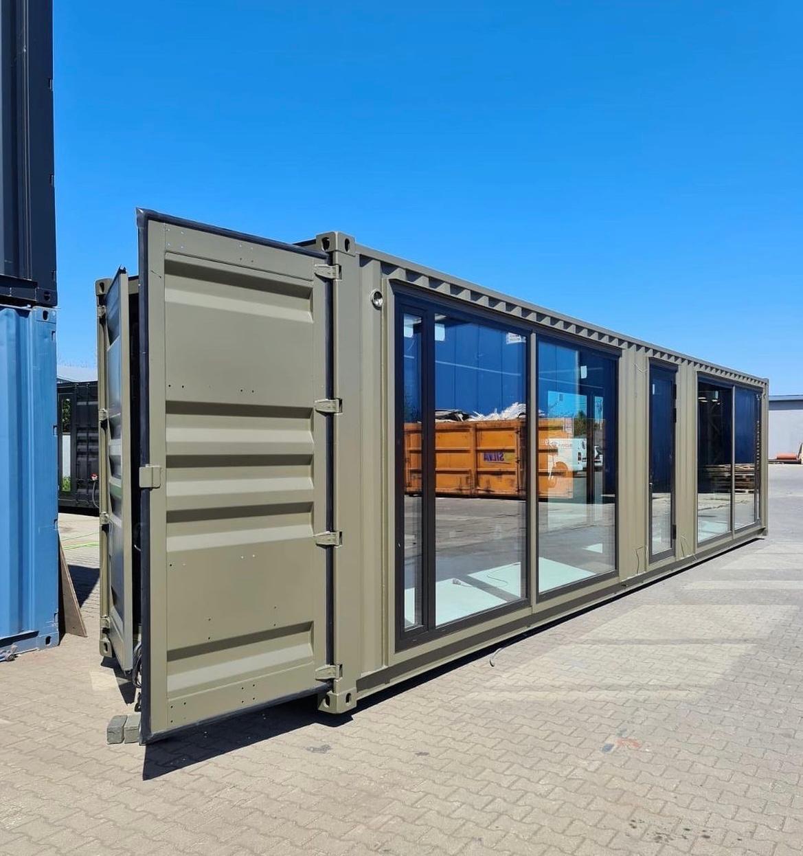 Ein grauer Wohncontainer mit einer großen Schiebetür in schwarz, sowie mehreren großen Fenstern in schwarz an der Frontseite. Der Wohn Container befindet sich im Freien.